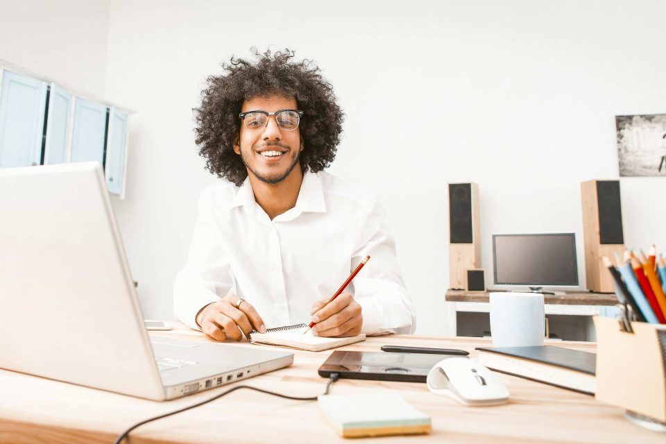 Curly-haired person at desk, pencil in hand, editing content on laptop, smiling.