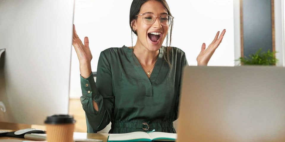 Person with glasses smiling, raising hands at a laptop: make your website stand out.