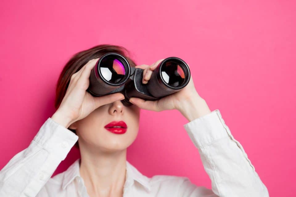 Person in white shirt looking through binoculars against pink background.