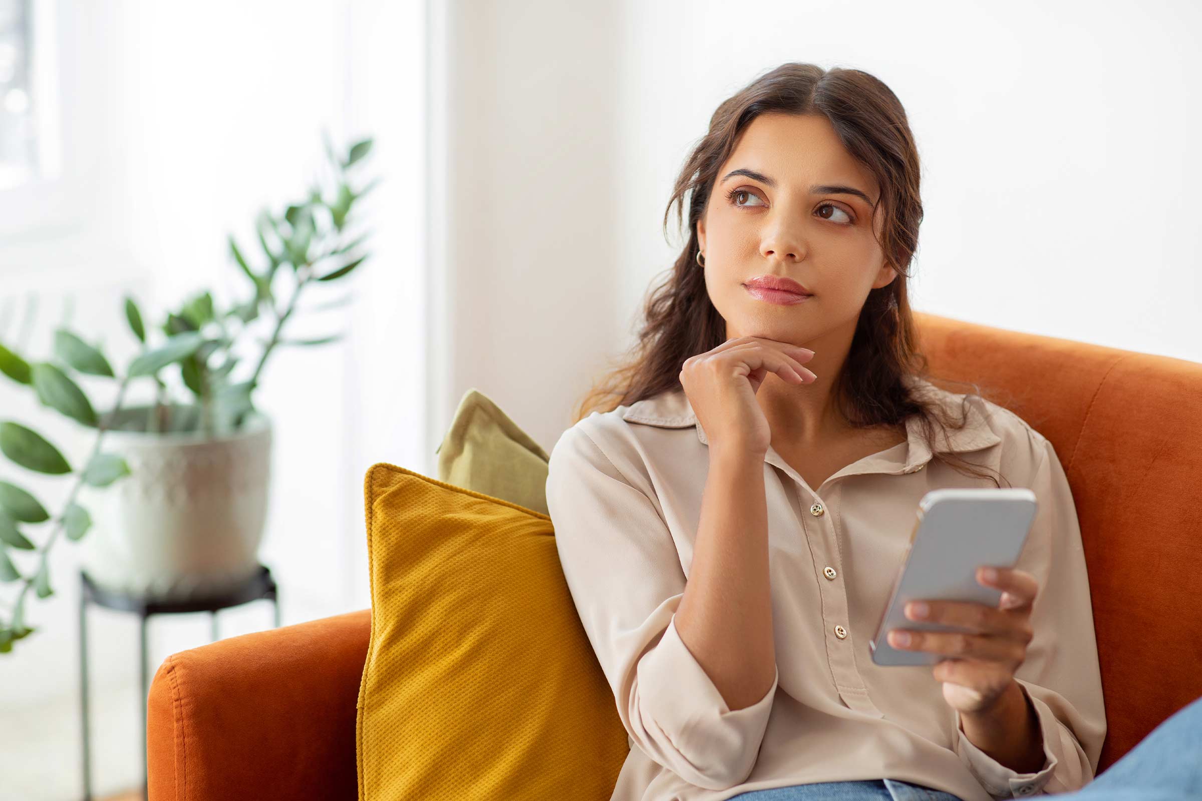 Woman on orange sofa with smartphone, pondering social media comment moderation.