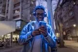 Person wearing headphones, smiling while looking at a smartphone outdoors.