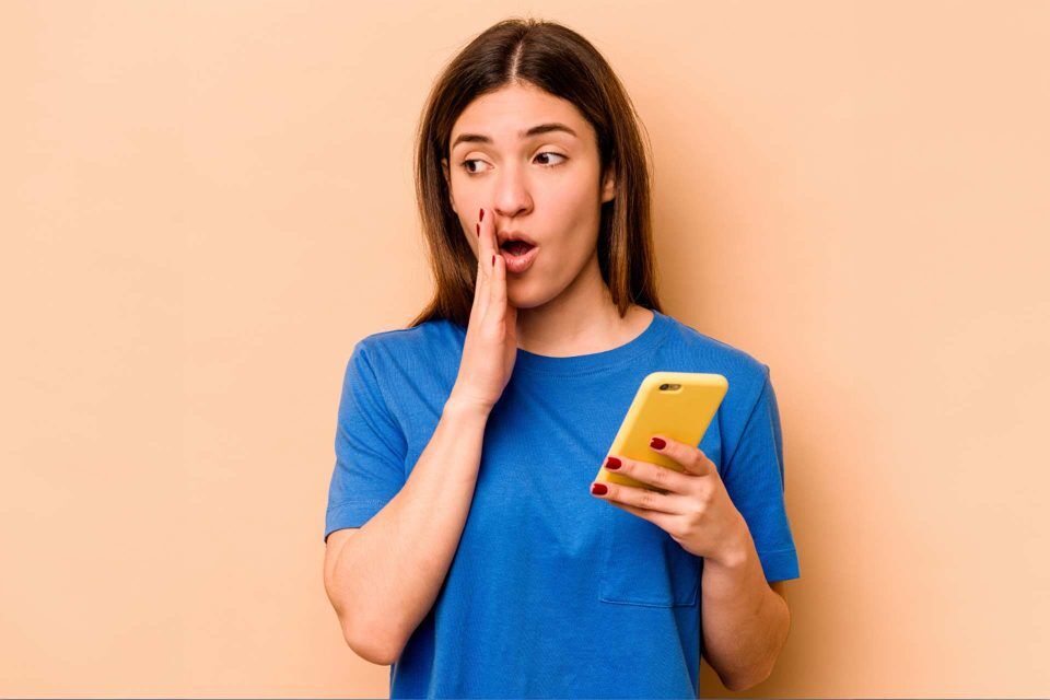 Woman in blue shirt, shocked by vanity metrics, holding yellow phone, beige backdrop.