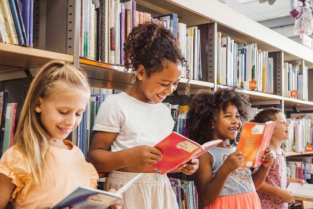 Children of Baker Youth Club read books in a library aisle, smiling in a row.