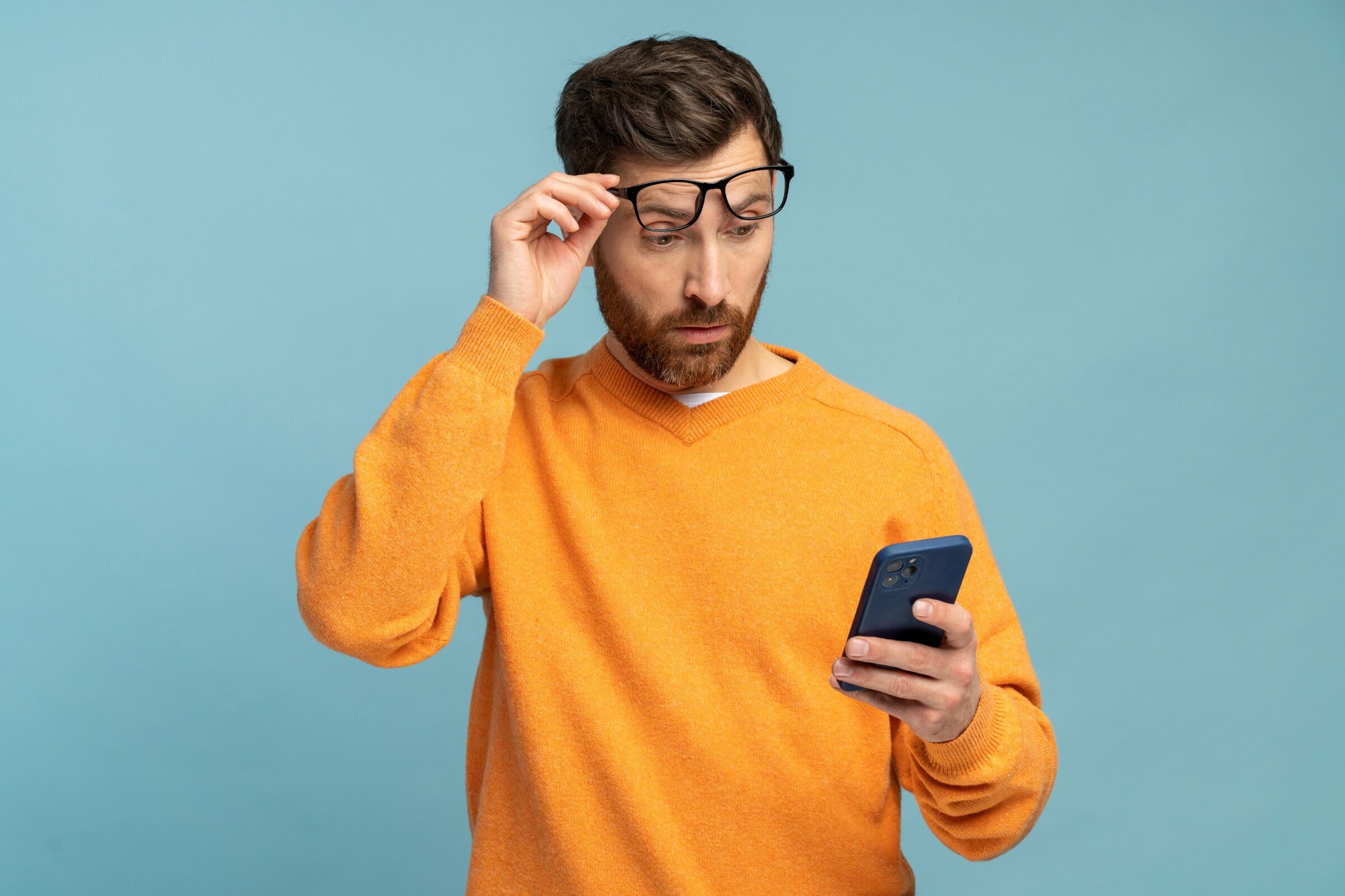 Man in orange sweater looks surprised at his phone, adjusting glasses.