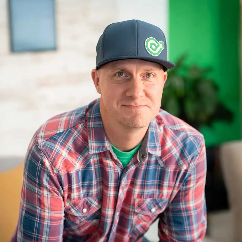 Man in plaid shirt and cap smiling at the camera indoors.