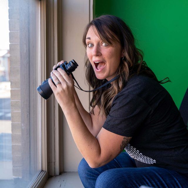 Person smiling, holding binoculars while kneeling by a window.