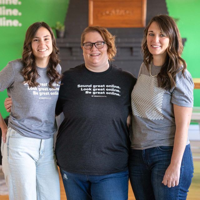 Three people standing indoors, smiling, in casual outfits.