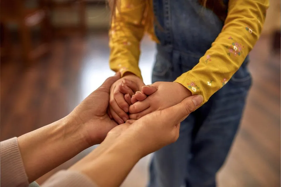 Adult holding hands with a child wearing a yellow long-sleeve shirt and overalls.
