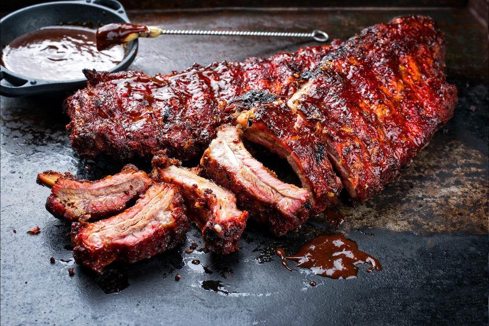 A rack of barbecued ribs with sauce and several ribs sliced on a dark tray.