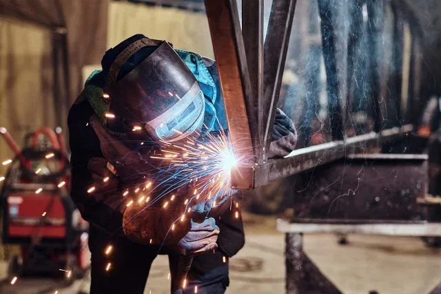 A person welding metal beams with sparks flying in an industrial workshop.