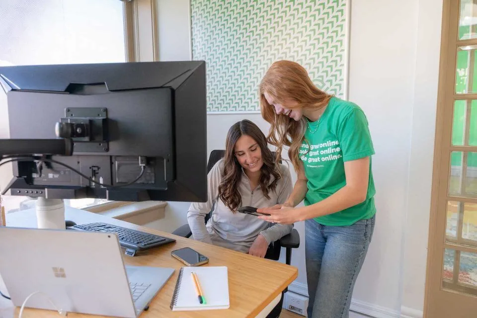 Two women in an office look at a phone together by a desk with computers.