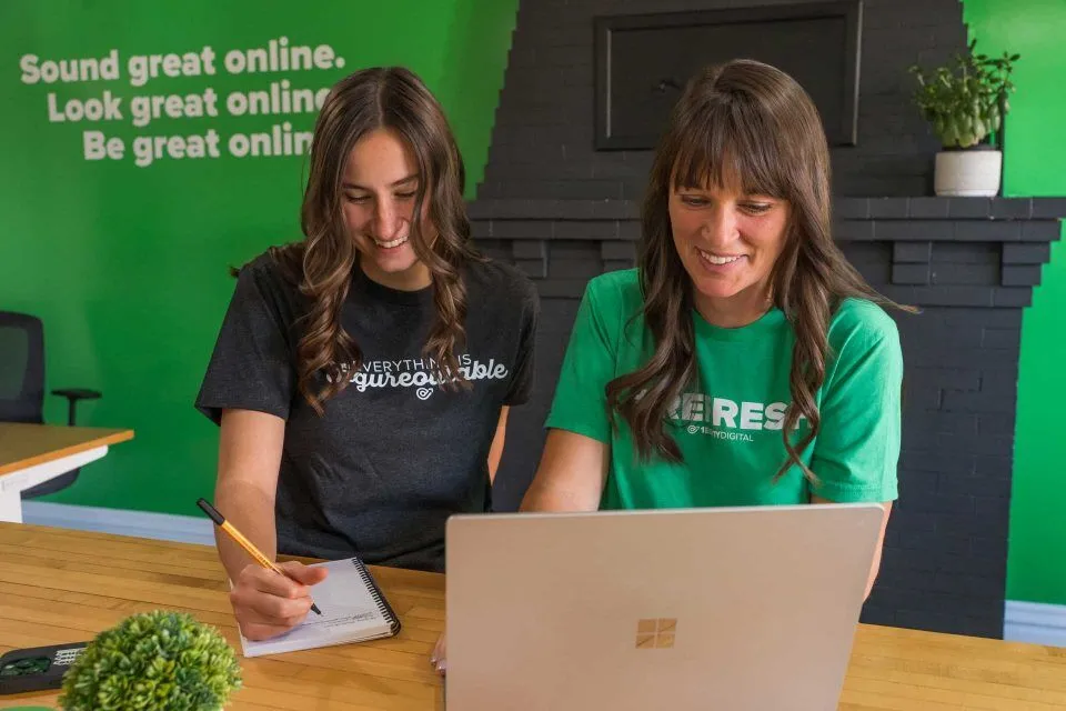 Two women working together at a laptop and taking notes in a green office.