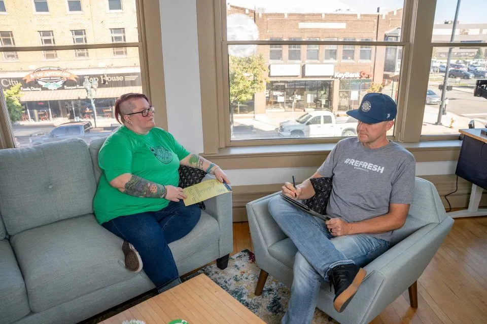Two people sit on couches in an office, talking and taking notes on clipboards.