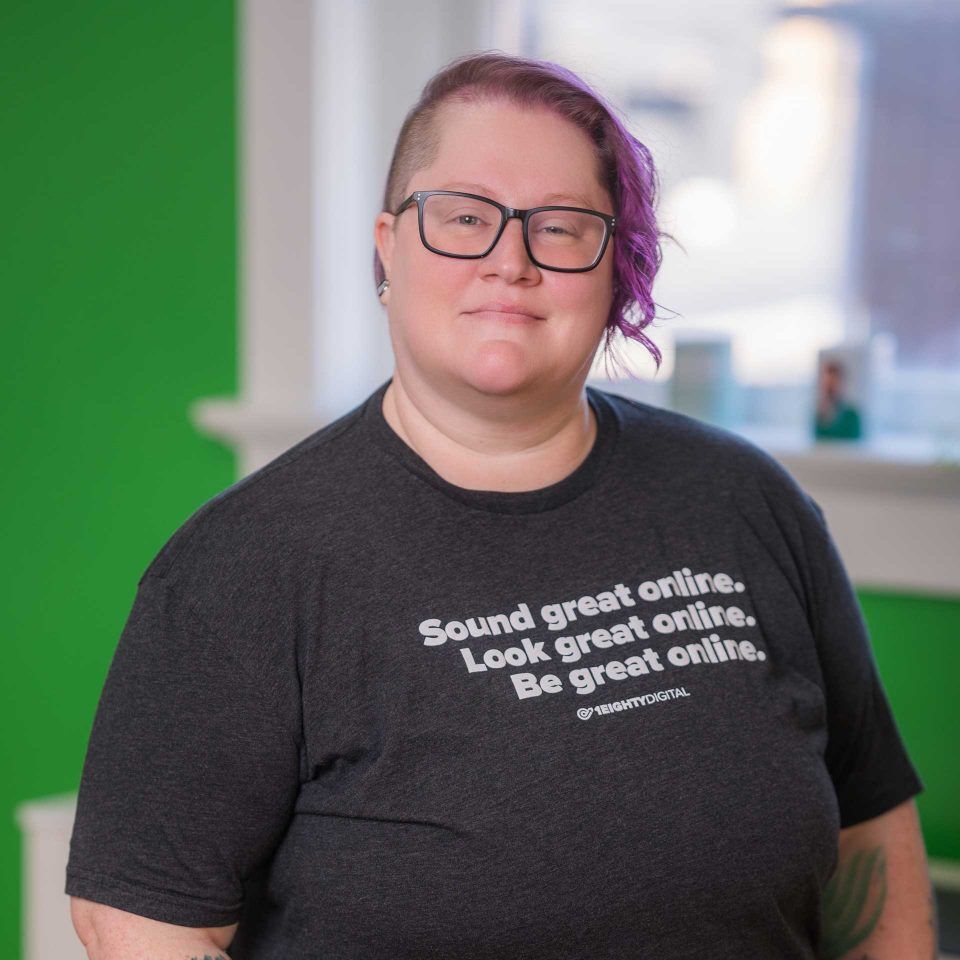 Person with short purple hair and glasses, wearing a graphic t-shirt, indoors.