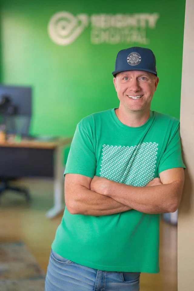 Man in green shirt and blue cap smiles, standing with arms crossed in office.