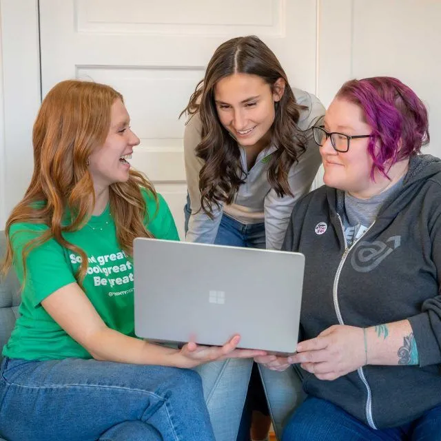 Three women sit together, smiling and looking at a laptop screen.
