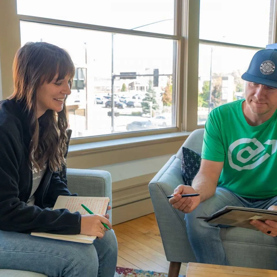 Two people sit on chairs, discussing notes and a tablet in a bright office.