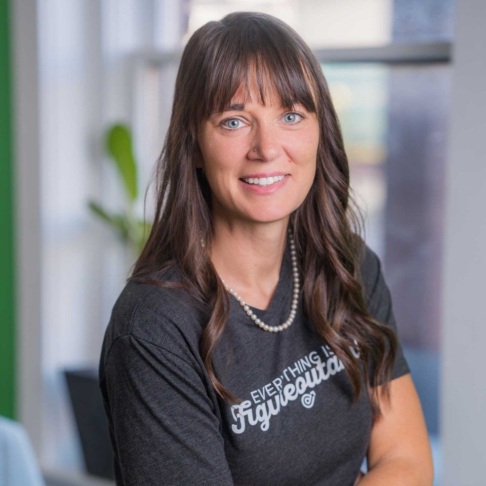Woman with long brown hair in a t-shirt, smiling indoors near a window.