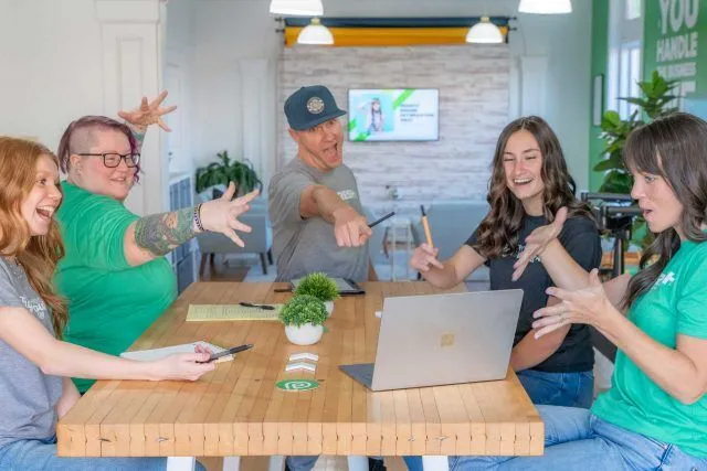 Five people sit around a table, smiling and gesturing excitedly at a laptop.