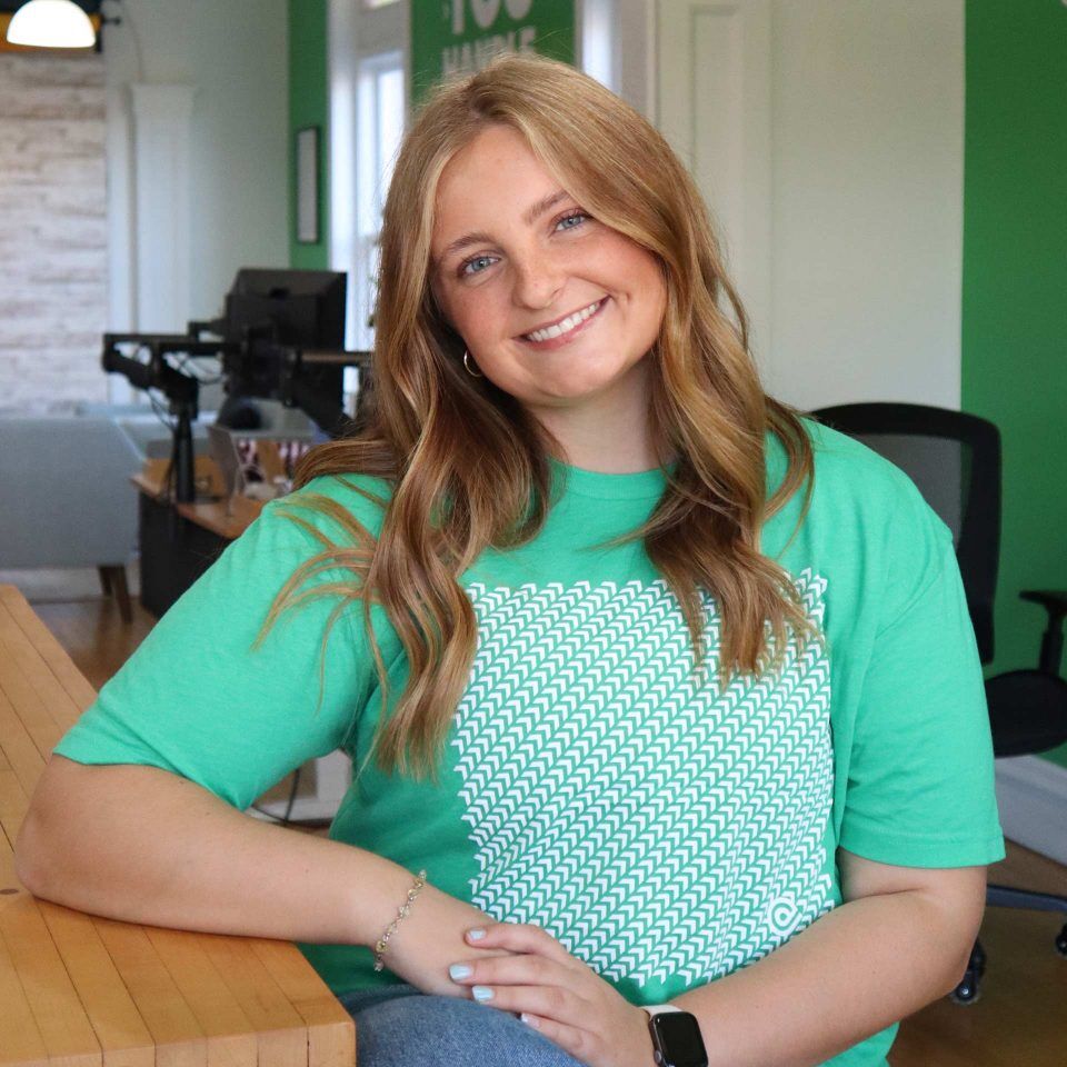 Woman in green t-shirt sits at a desk, smiling in a bright office setting.