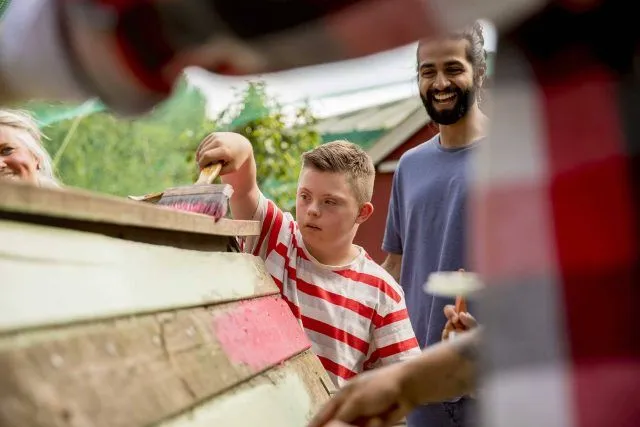 Child painting a wooden structure with adults supervising outdoors.