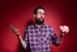 Man holding a smartphone and piggy bank, pondering his marketing budget.