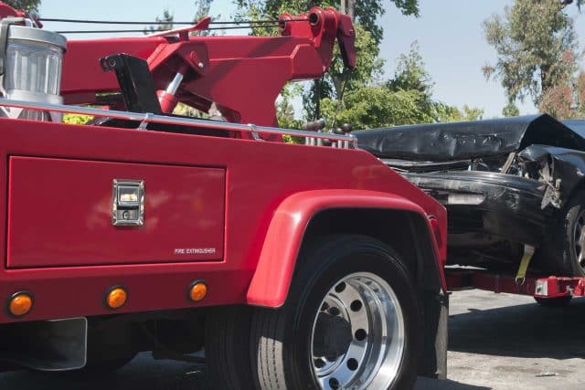 Tow truck from Crouse Body Shop towing a damaged black car in the sun.