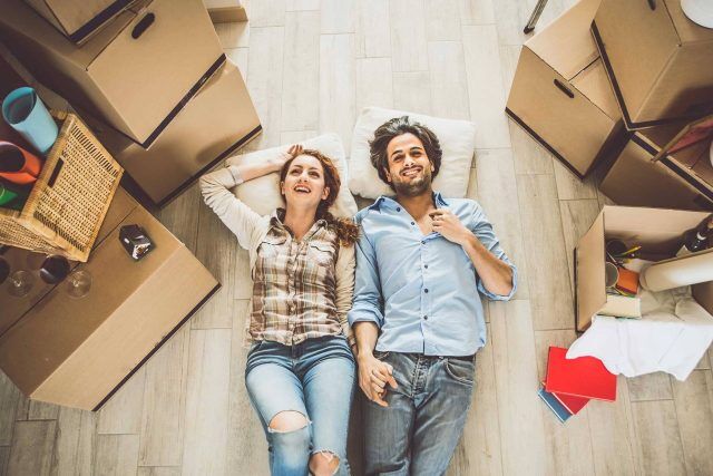 Couple lying on floor among moving boxes, smiling and holding hands.