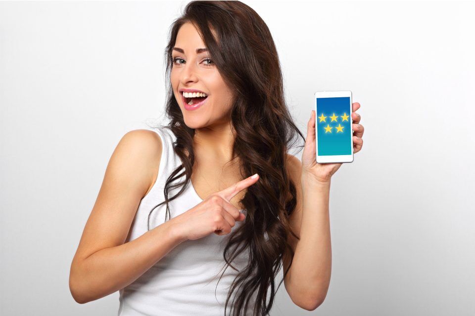 Smiling woman points at phone showcasing 5-star reviews.