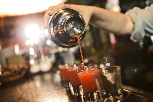 Bartender pouring red cocktails into shot glasses at a bar.