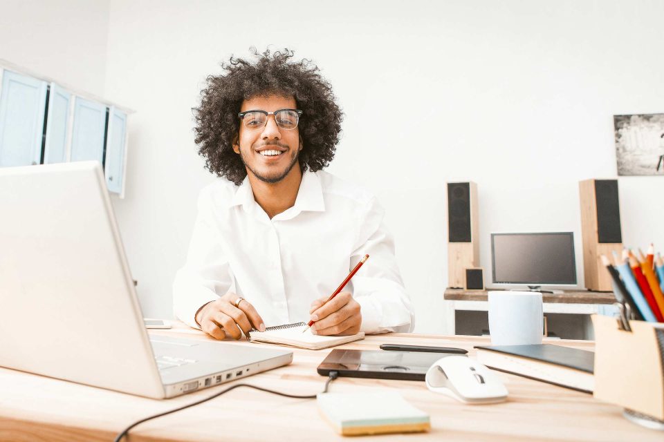 Curly-haired person at desk, pencil in hand, editing content on laptop, smiling.