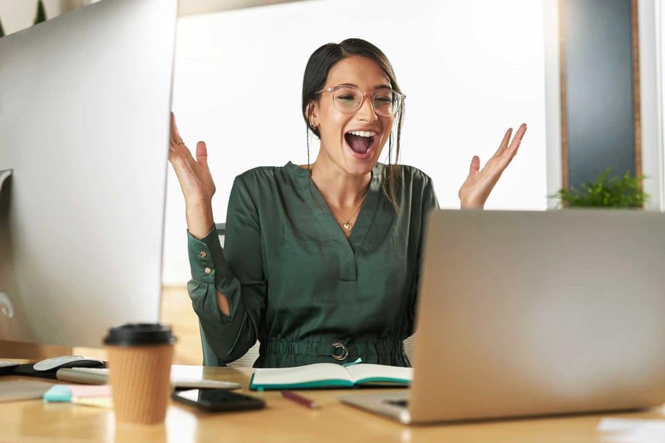 Person with glasses smiling, raising hands at a laptop: make your website stand out.