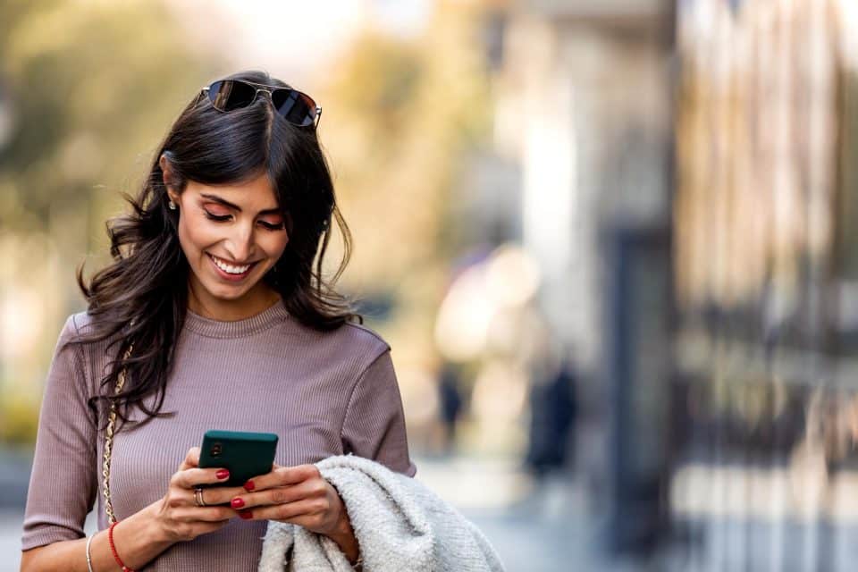 Woman smiling at phone while walking, checking Facebook ads, holding a jacket.