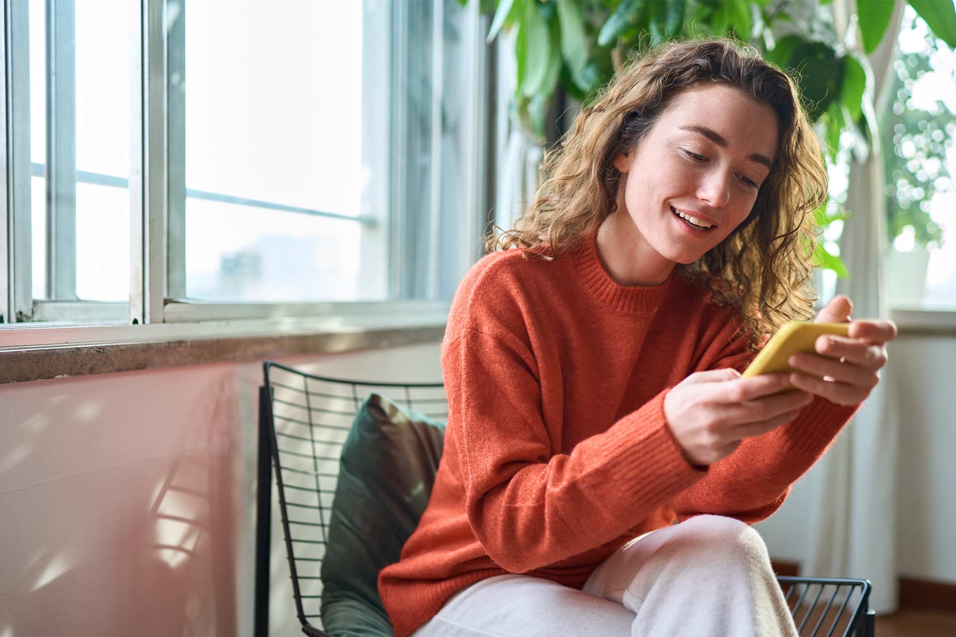 Woman sitting by a window, smiling at clever email subject lines on her phone.