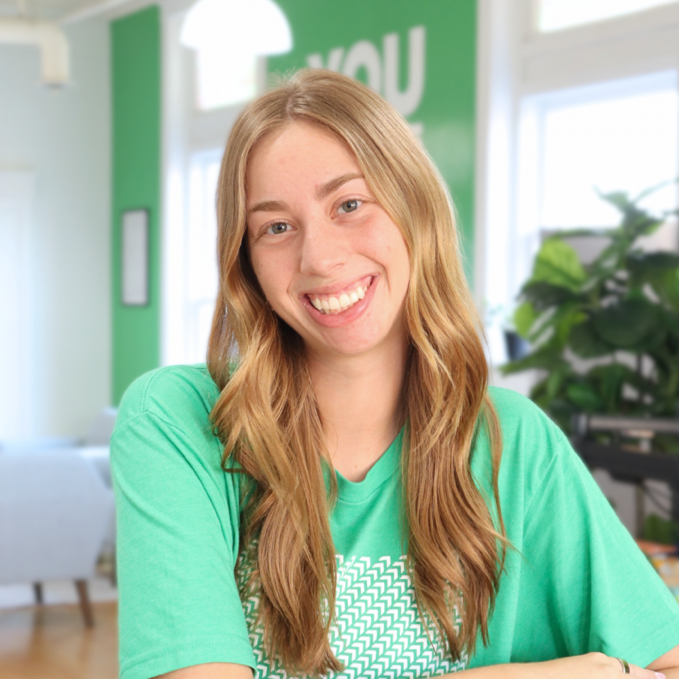 Woman with long blonde hair smiling, wearing a green shirt in a bright room.