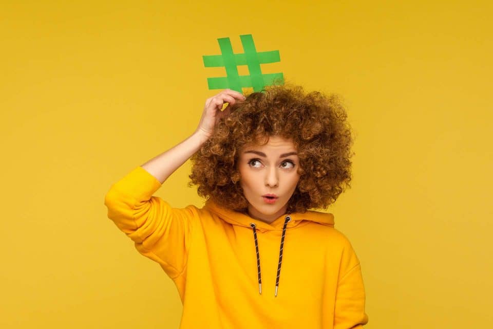 Person wearing a yellow hoodie, holding a green hashtag sign on head.