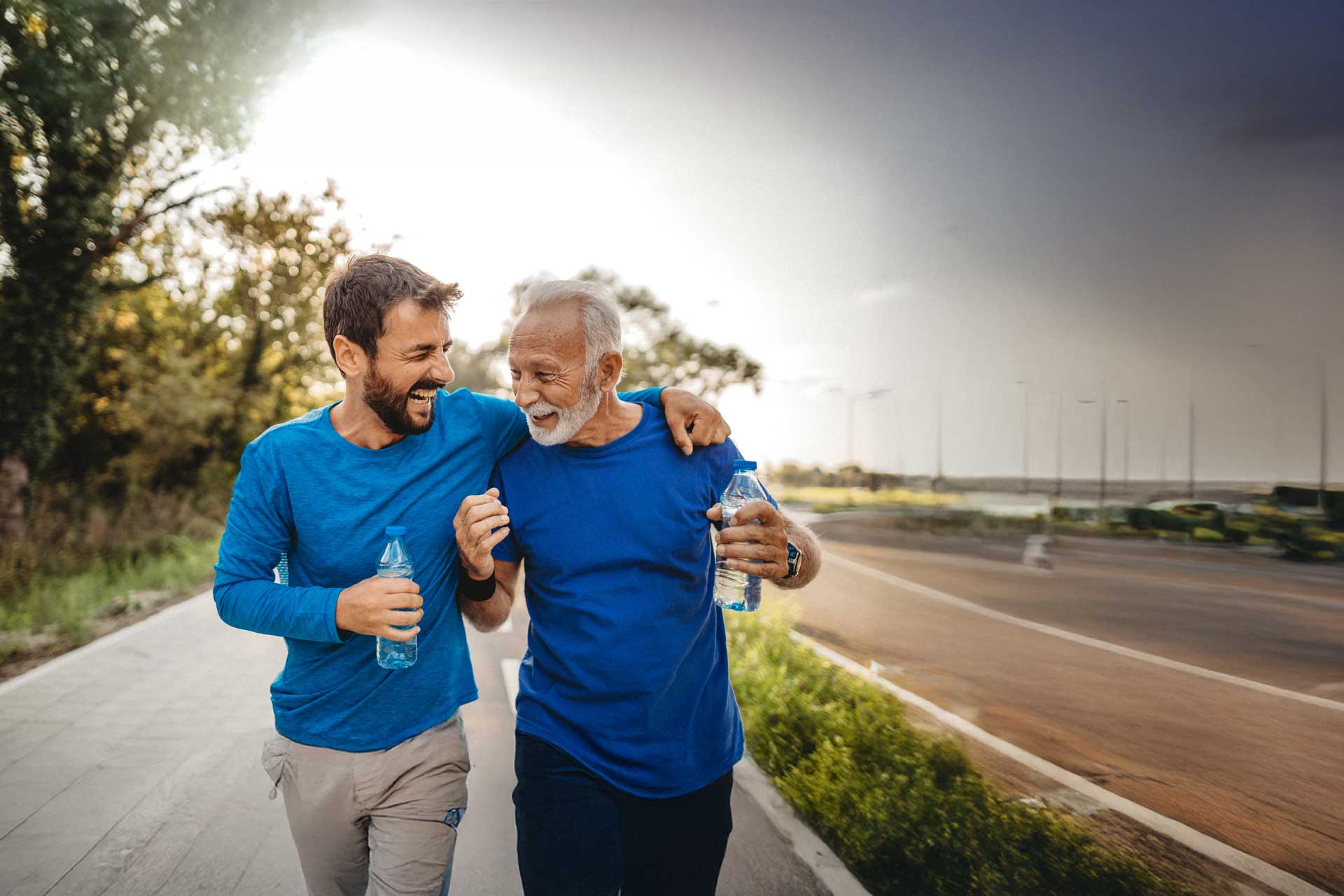 Two men walking outdoors with water bottles, smiling and embracing.
