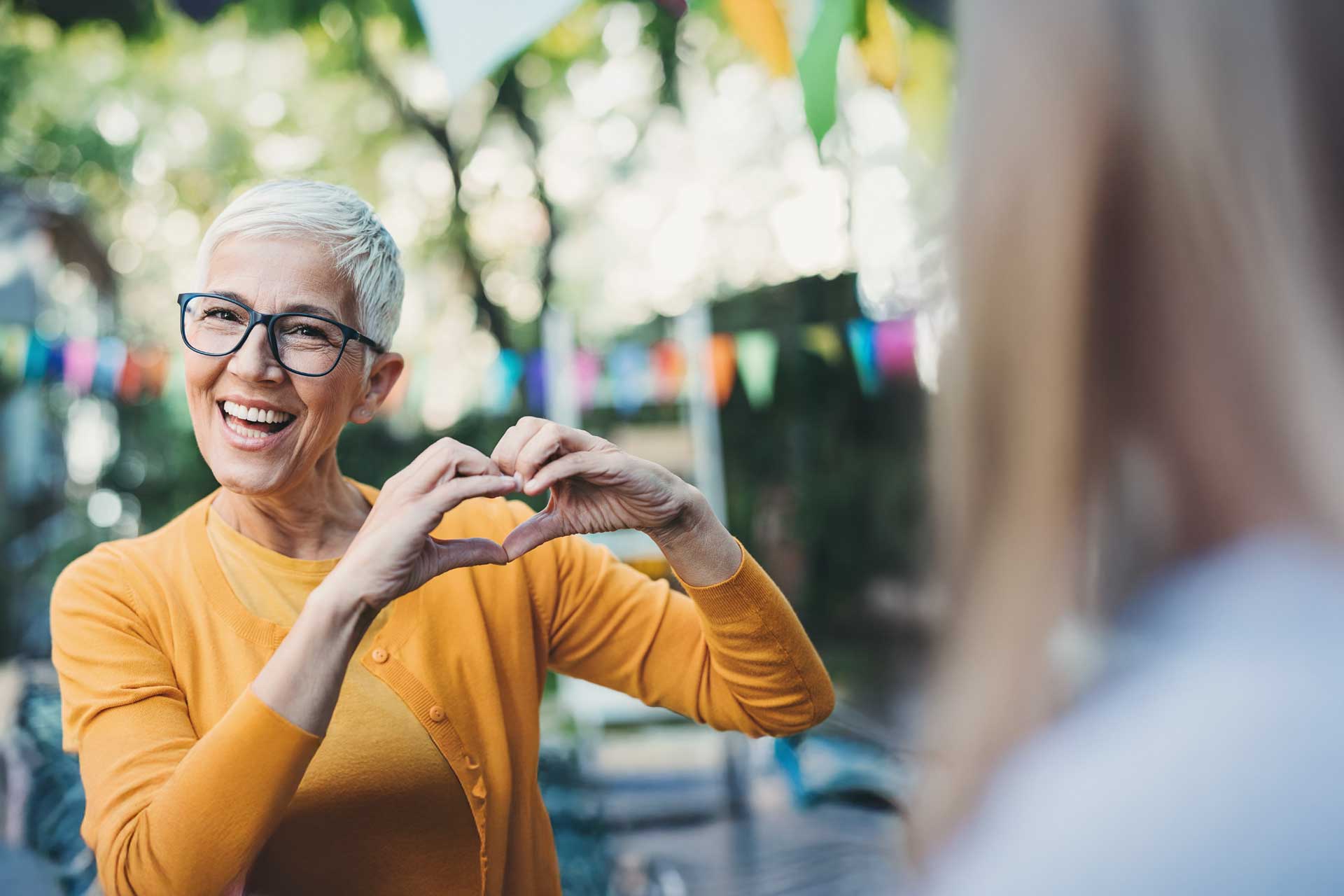 Smiling woman in yellow makes heart gesture outdoors near colorful bunting.