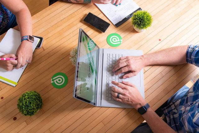 Two people working on a laptop at a wooden table, with notebooks and plants.