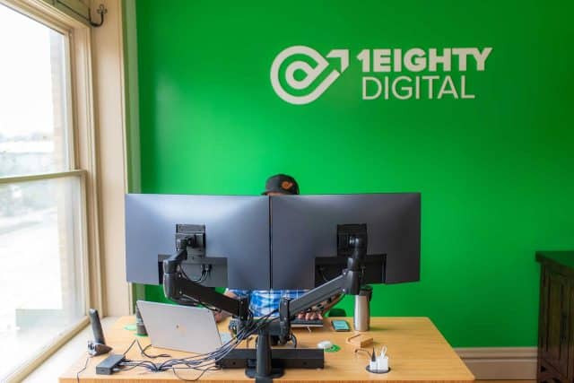 Person working at dual monitors in a green office with 1EIGHTY DIGITAL logo.