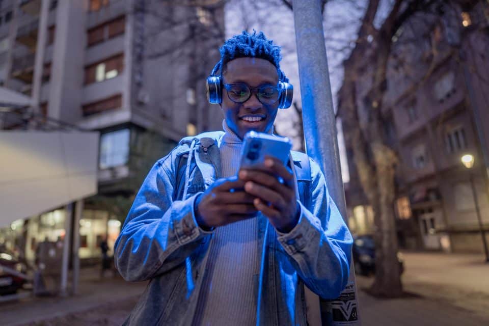 Person wearing headphones, smiling while looking at a smartphone outdoors.