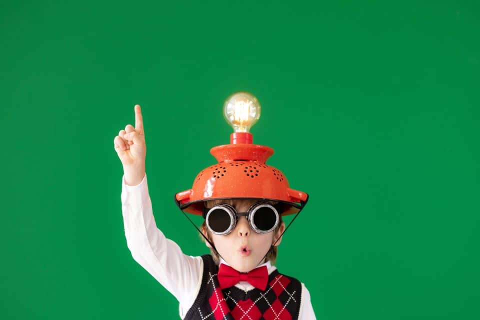 Child with lightbulb helmet and goggles raising finger, green background.