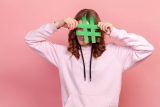 Person in pink hoodie hides behind a sizeable green hashtag for research.