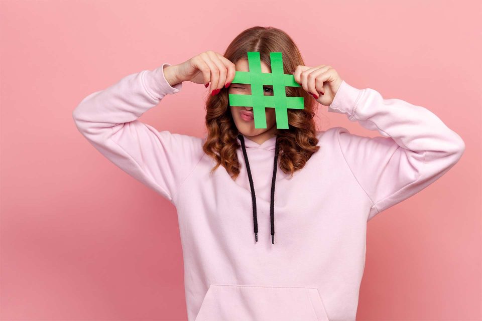Person in pink hoodie hides behind a sizeable green hashtag for research.