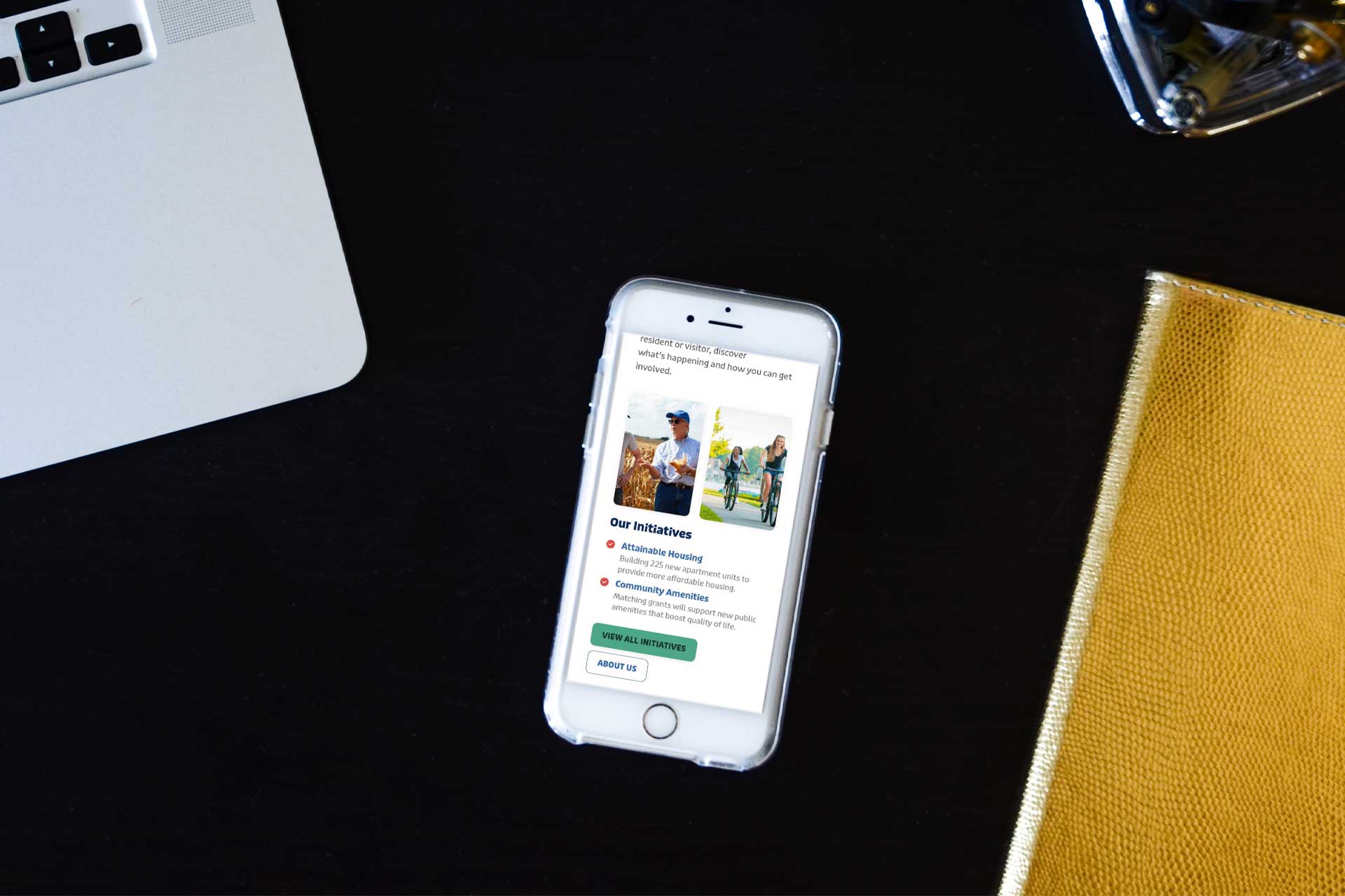 A smartphone on a black desk displays a webpage next to a laptop and yellow notebook.