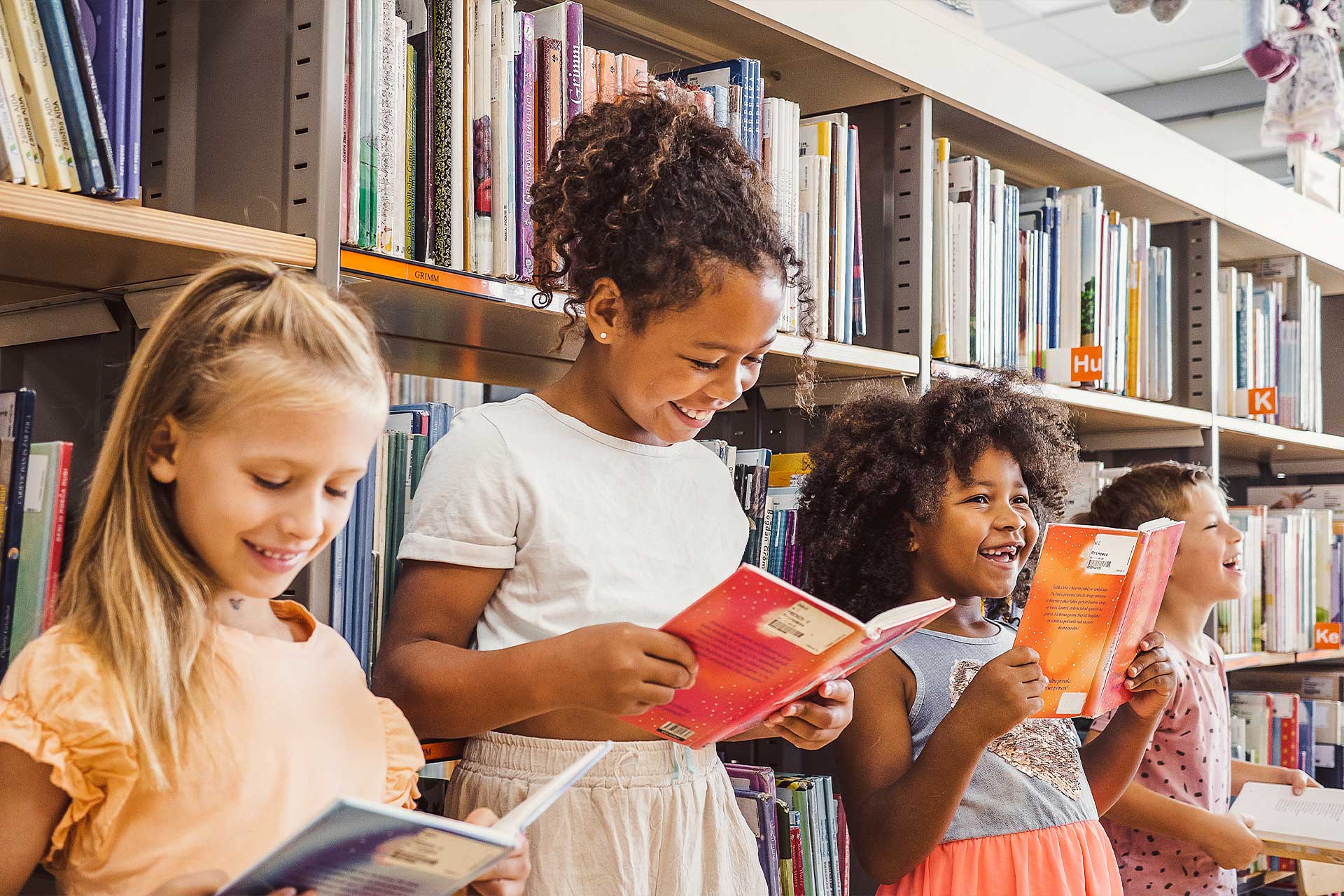 Children of Baker Youth Club read books in a library aisle, smiling in a row.