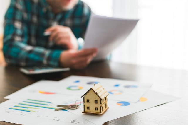 Small house model and keys on papers with charts; person reviews documents in background.