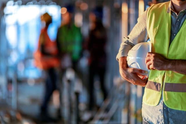 Person in safety vest holding hard hat at a construction site with workers in background.