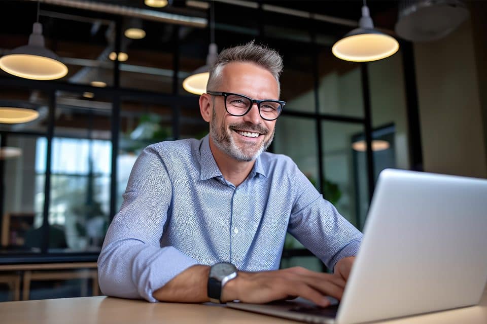 Man with glasses smiling while working on a laptop in a modern office.