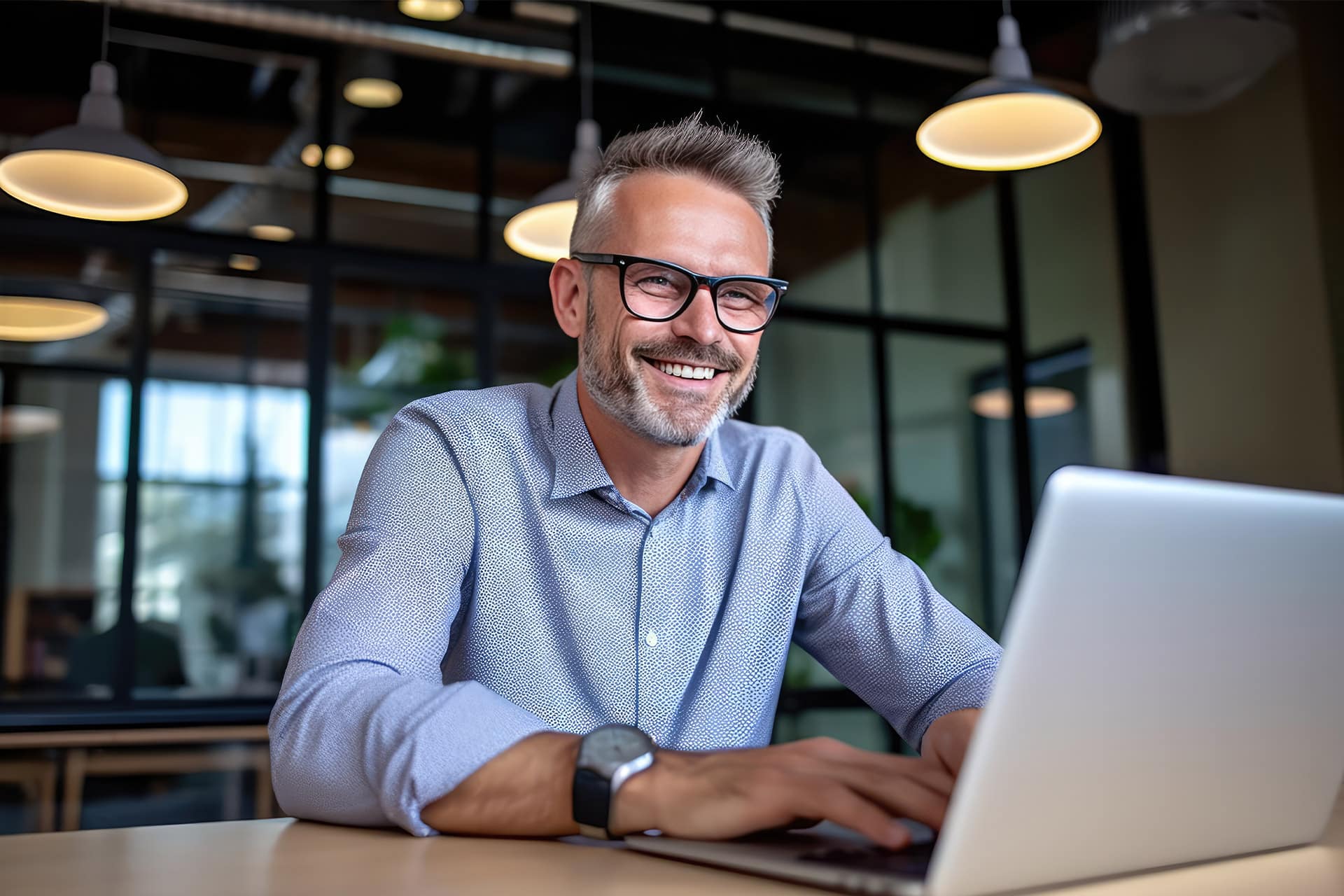 Man with glasses smiling while working on a laptop in a modern office.